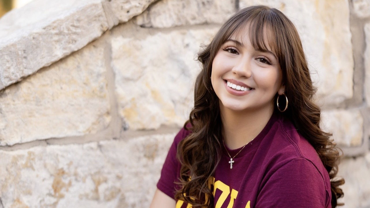 Olivia Tomasino smiles in front of a stone wall while wearing a maroon t-shirt Olivia Tomasino smiles in front of a stone wall while wearing a maroon t-shirt
